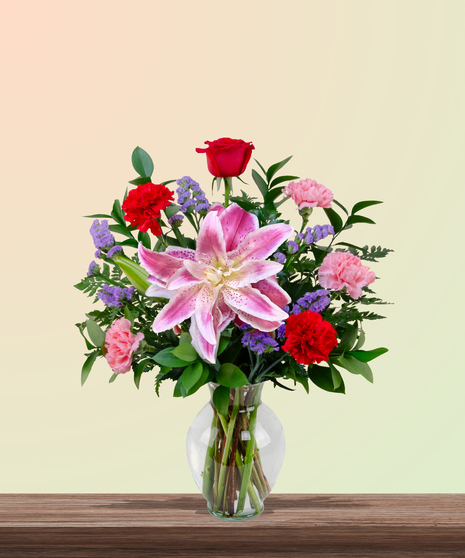 Mother’s Touch floral arrangement with red roses, pink carnations, stargazer lilies, and purple statice in a clear glass vase – a classic Mother’s Day gift.