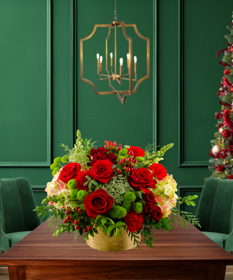 Noel classic Christmas flower arrangement with red roses, red carnations, green button mums, green hydrangeas, Bells of Ireland, and evergreen in a gold textured container