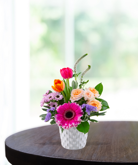 Radiant Energy floral arrangement with hot pink gerbera daisy, orange tulips, peach spray roses, and white veronica in a white textured honeycomb vase.
