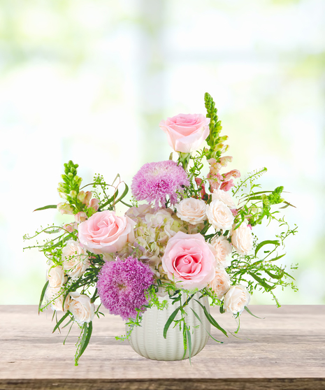 Lavender Sunrise floral arrangement with peach and blush roses, lavender pom blooms, snapdragons, and hydrangeas in a white ceramic vase.