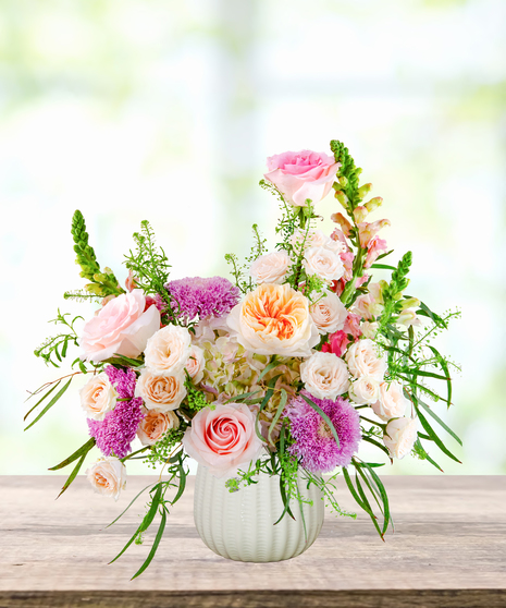 Lavender Sunrise floral arrangement with peach and blush roses, lavender pom blooms, snapdragons, and hydrangeas in a white ceramic vase.