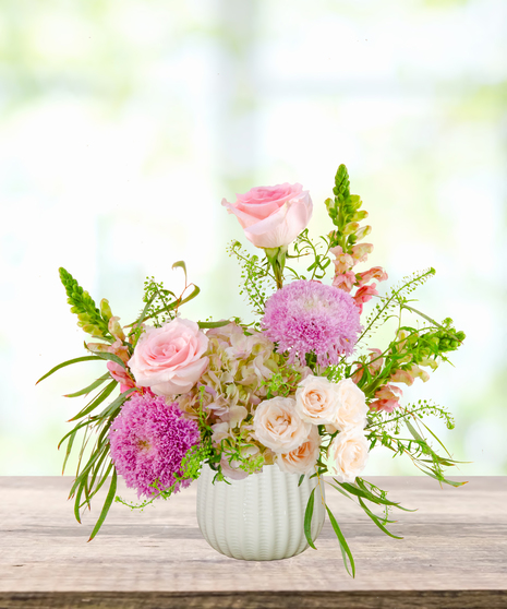 Lavender Sunrise floral arrangement with peach and blush roses, lavender pom blooms, snapdragons, and hydrangeas in a white ceramic vase.