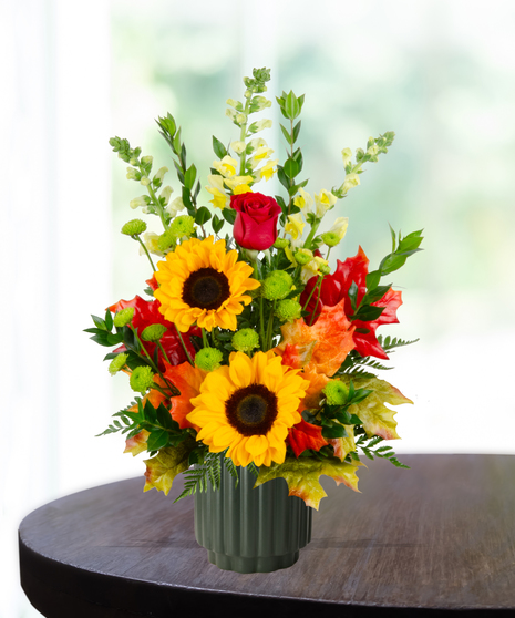 Orchard Sunset flower arrangement with sunflowers, red roses, yellow snapdragons, and autumn leaves in a green vase.