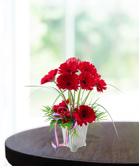 Party in the USA patriotic flower arrangement featuring bright red gerbera daisies, red berries, lily grass, and a red, white, and blue ribbon in a white textured vase.