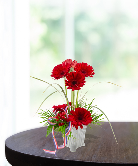 Party in the USA patriotic flower arrangement featuring bright red gerbera daisies, red berries, lily grass, and a red, white, and blue ribbon in a white textured vase.