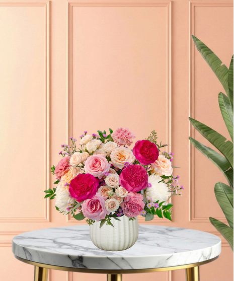 Peepin’ Pretty floral arrangement with blush and fuchsia roses, carnations, ivory mums, and lavender asters in a white ceramic vase.