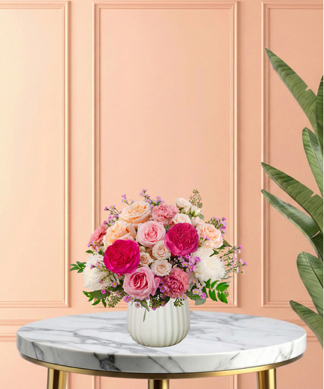 Peepin’ Pretty floral arrangement with blush and fuchsia roses, carnations, ivory mums, and lavender asters in a white ceramic vase.