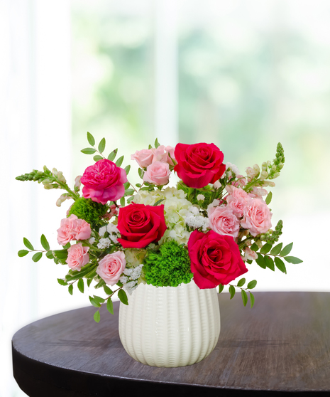 She’s All That! floral arrangement with hot pink roses, blush spray roses, green hydrangeas, snapdragons, and green dianthus in a white pedestal vase.