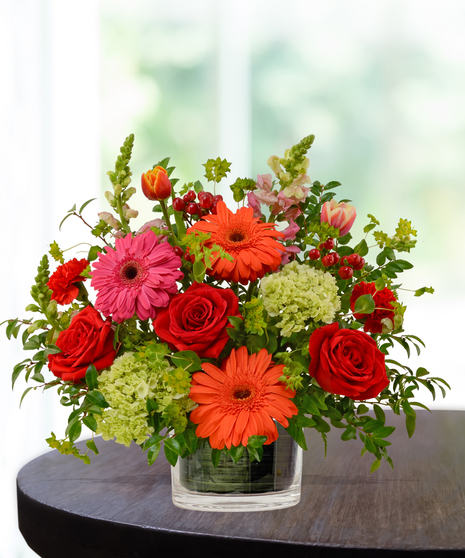 Heatwave flower arrangement with red roses, orange gerberas, pink daisies, and pincushion protea in a clear glass cube vase