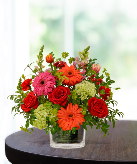 Heatwave flower arrangement with red roses, orange gerberas, pink daisies, and pincushion protea in a clear glass cube vase