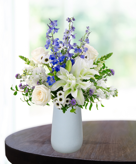 September birthday flower arrangement with blue hydrangeas, white roses, lilies, and delphinium in a soft blue vase.