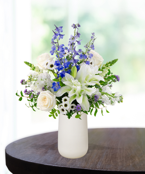 December Skies birthday flower arrangement with white roses, white lilies, blue delphinium, blue hydrangeas, white snapdragons, and green foliage in a frosted white vase
