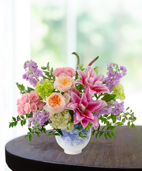 Splash of Spring March birthday arrangement featuring pink lilies, peach roses, purple stock, green hydrangeas, and tall veronica in a blue and white marbled ceramic vase