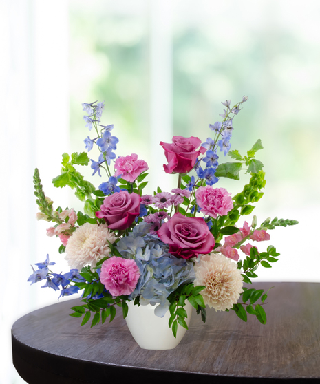 Malibu Splash flower arrangement featuring magenta garden roses, lavender roses, blue hydrangeas, and blue delphinium in a matte white ceramic bowl.