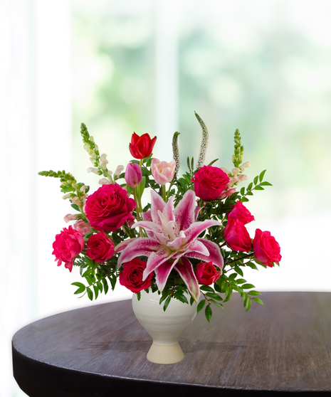 Legally Pink flower arrangement featuring hot pink roses, pink lilies, pink tulips, and white veronica in a matte cream ceramic pedestal vase.