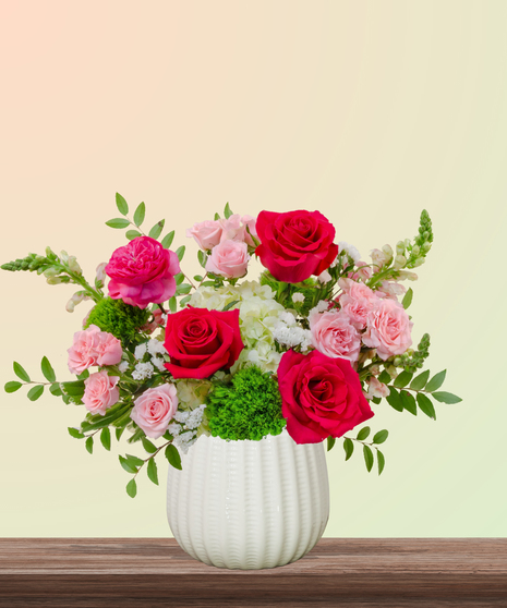 She’s All That! floral arrangement with hot pink roses, blush spray roses, green hydrangeas, snapdragons, and green dianthus in a white pedestal vase.