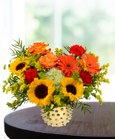 Bright fall flower arrangement with sunflowers, orange roses, red carnations, and gerbera daisies in a gold vase