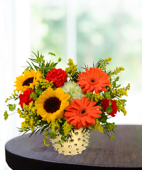 Bright fall flower arrangement with sunflowers, orange roses, red carnations, and gerbera daisies in a gold vase