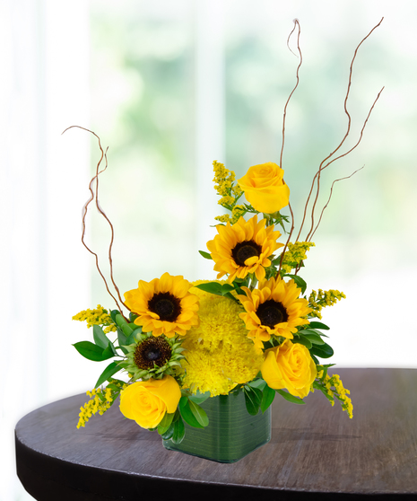 Bright sunflower and yellow rose flower arrangement in a leaf wrapped cube vase.
