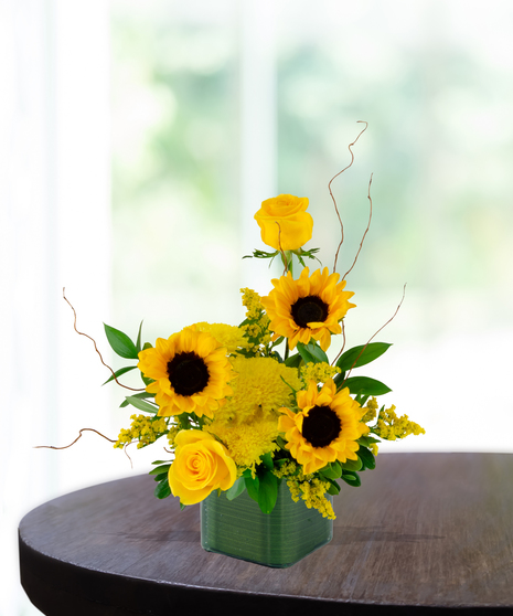 Bright sunflower and yellow rose flower arrangement in a leaf wrapped cube vase.