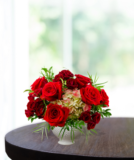 True Love anniversary floral arrangement featuring premium red roses, red spray roses, and lime-green hydrangeas in a white textured beaded pedestal vase.