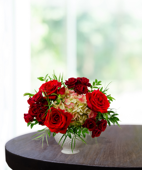 True Love anniversary floral arrangement featuring premium red roses, red spray roses, and lime-green hydrangeas in a white textured beaded pedestal vase.