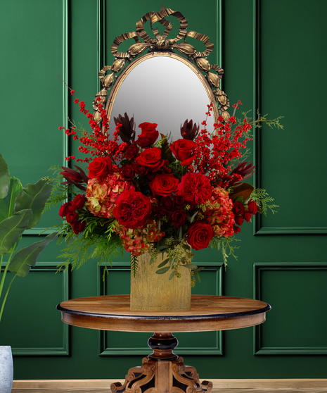 Rouge luxury Christmas flower arrangement with deep red roses, red hypericum berries, dark red foliage, and green accents in a gold vase