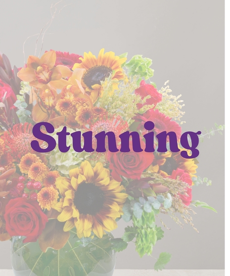 Saffron flower arrangement with sunflowers, cymbidium orchids, red roses, protea, and fall accents in a round green glass vase.