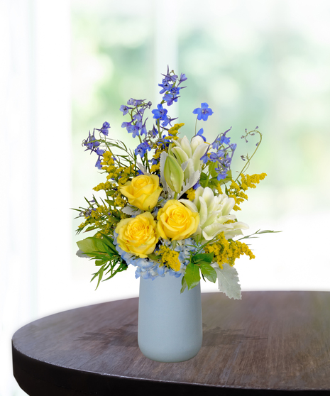 Spirit of the Bluff collegiate flower arrangement featuring yellow roses, white lilies, blue hydrangeas, and blue delphinium in a light blue matte vase.