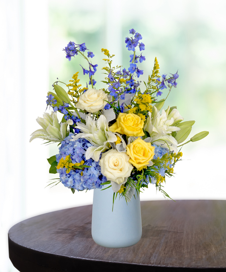 Spirit of the Bluff collegiate flower arrangement featuring yellow roses, white lilies, blue hydrangeas, and blue delphinium in a light blue matte vase.