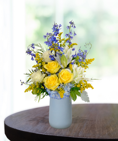 Spirit of the Bluff collegiate flower arrangement featuring yellow roses, white lilies, blue hydrangeas, and blue delphinium in a light blue matte vase.