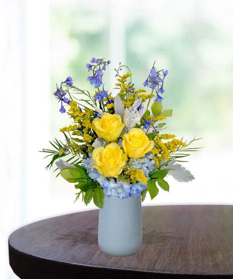 Spirit of the Bluff collegiate flower arrangement featuring yellow roses, white lilies, blue hydrangeas, and blue delphinium in a light blue matte vase.