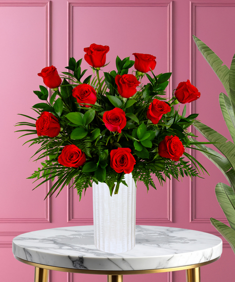 A romantic floral arrangement featuring red roses and lush green foliage, displayed in a sleek white vase against a pink background.
