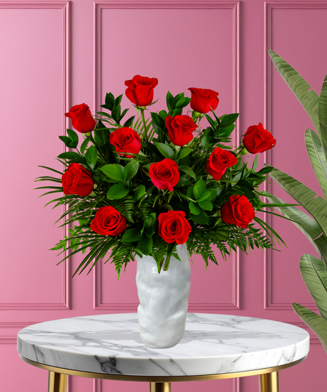 A romantic floral arrangement featuring red roses and lush green foliage, displayed in a sleek white vase against a pink background.