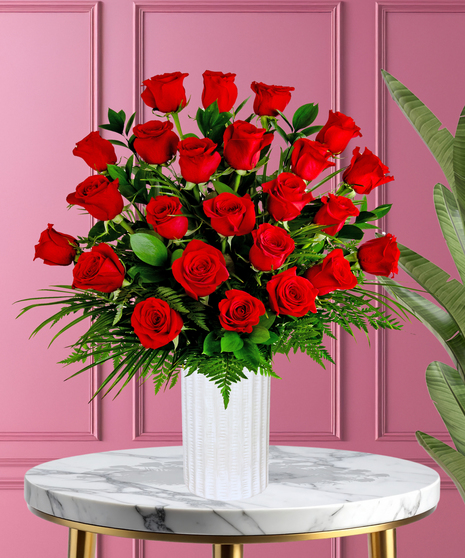A romantic floral arrangement featuring red roses and lush green foliage, displayed in a sleek white vase against a pink background.
