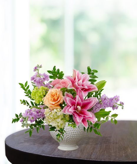 Splash of Spring March birthday arrangement featuring pink lilies, peach roses, purple stock, green hydrangeas, and tall veronica in a blue and white marbled ceramic vase