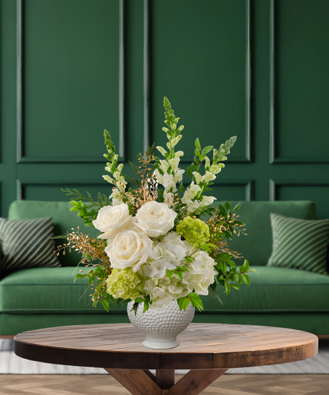 White Christmas flower arrangement with roses, hydrangeas, green hydrangeas, white snapdragons, evergreen, and gold/white sparkling accents in a textured white vase