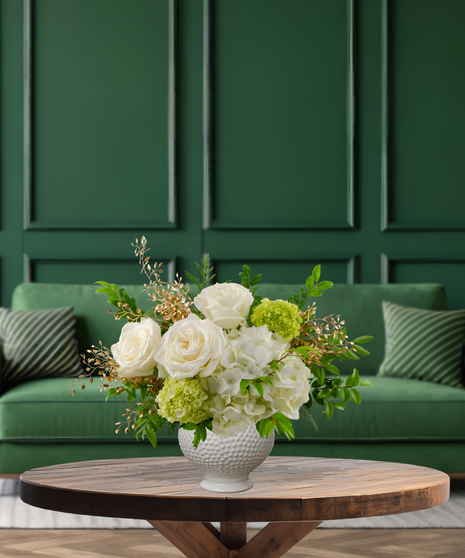 White Christmas flower arrangement with roses, hydrangeas, green hydrangeas, white snapdragons, evergreen, and gold/white sparkling accents in a textured white vase
