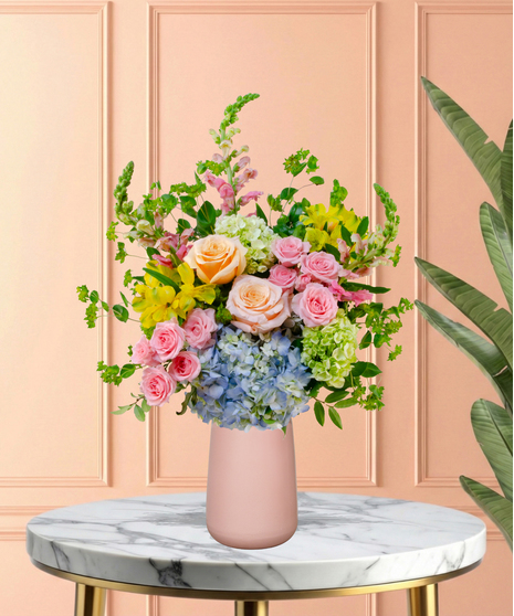 Spring Sorbet Easter flower arrangement with roses, spray roses, blue and green hydrangeas, alstroemeria, snapdragons, and lush green foliage in a matte blush-pink vase