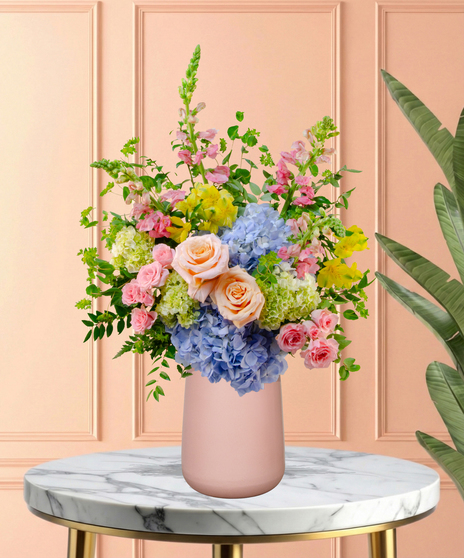 Spring Sorbet Easter flower arrangement with roses, spray roses, blue and green hydrangeas, alstroemeria, snapdragons, and lush green foliage in a matte blush-pink vase