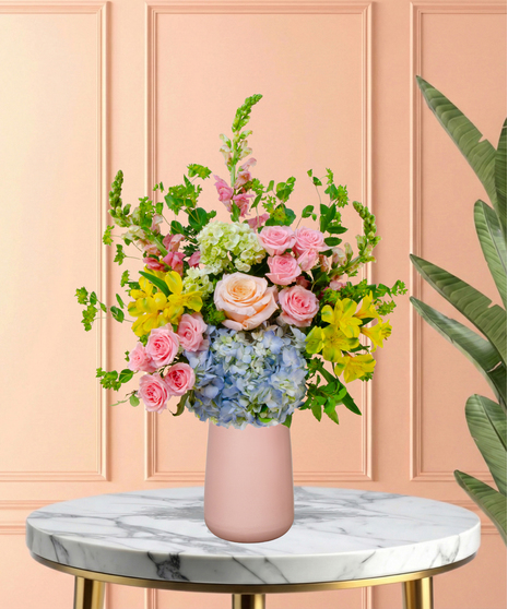 Spring Sorbet Easter flower arrangement with roses, spray roses, blue and green hydrangeas, alstroemeria, snapdragons, and lush green foliage in a matte blush-pink vase