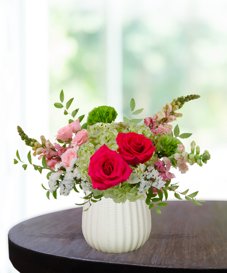 She’s All That! floral arrangement with hot pink roses, blush spray roses, green hydrangeas, snapdragons, and green dianthus in a white pedestal vase.