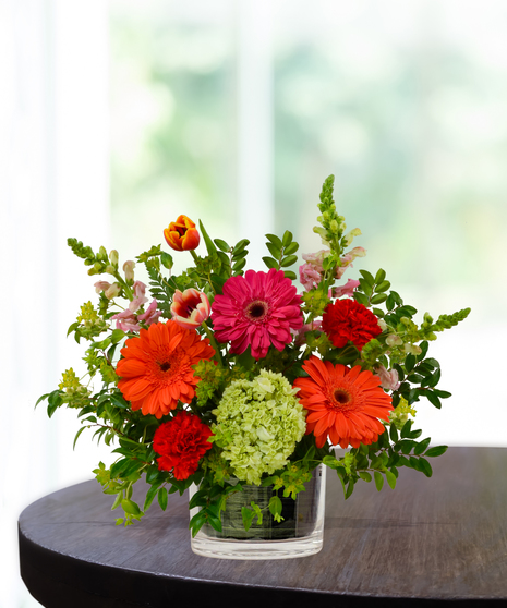 Heatwave flower arrangement with red roses, orange gerberas, pink daisies, and pincushion protea in a clear glass cube vase