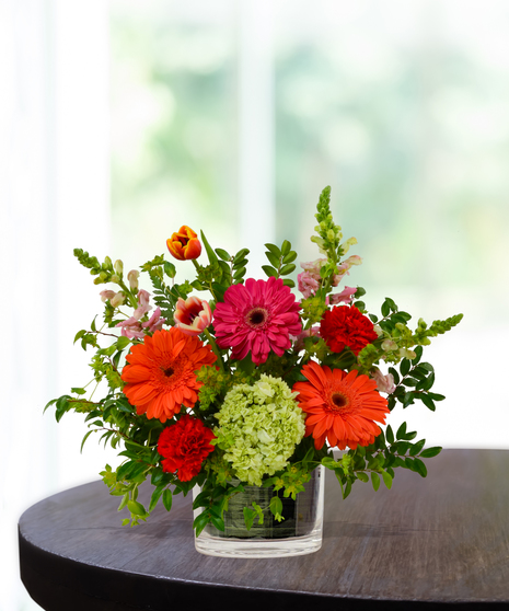 Heatwave flower arrangement with red roses, orange gerberas, pink daisies, and pincushion protea in a clear glass cube vase