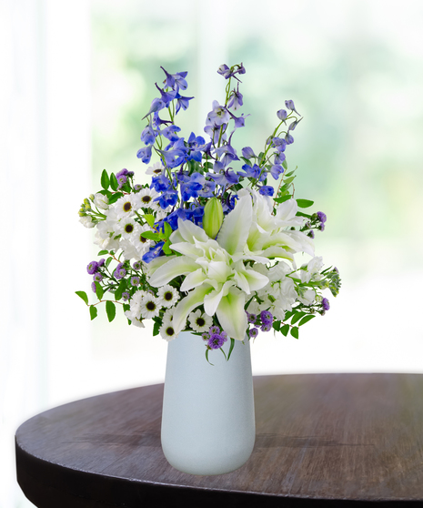 September birthday flower arrangement with blue hydrangeas, white roses, lilies, and delphinium in a soft blue vase.