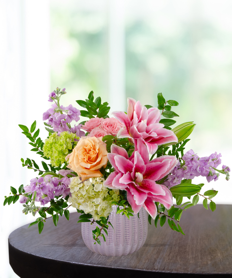 Spellbound flower arrangement with pink rose lilies, peach garden roses, pink spray roses, lavender stock, and green hydrangea in a soft blush vase
