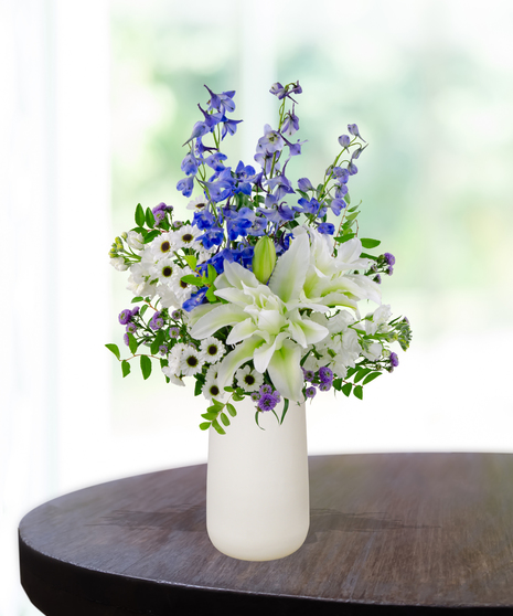 December Skies birthday flower arrangement with white roses, white lilies, blue delphinium, blue hydrangeas, white snapdragons, and green foliage in a frosted white vase