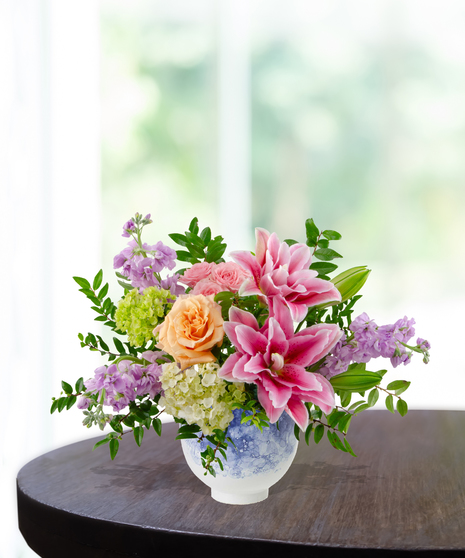 Splash of Spring March birthday arrangement featuring pink lilies, peach roses, purple stock, green hydrangeas, and tall veronica in a blue and white marbled ceramic vase
