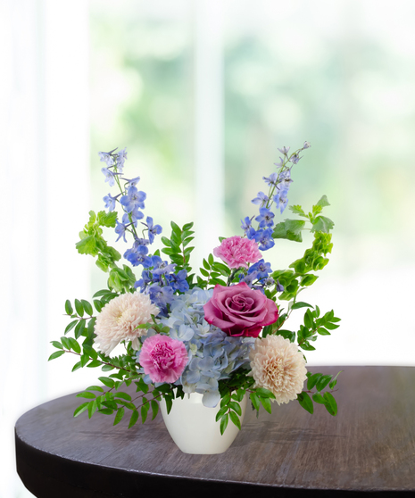 Malibu Splash flower arrangement featuring magenta garden roses, lavender roses, blue hydrangeas, and blue delphinium in a matte white ceramic bowl.