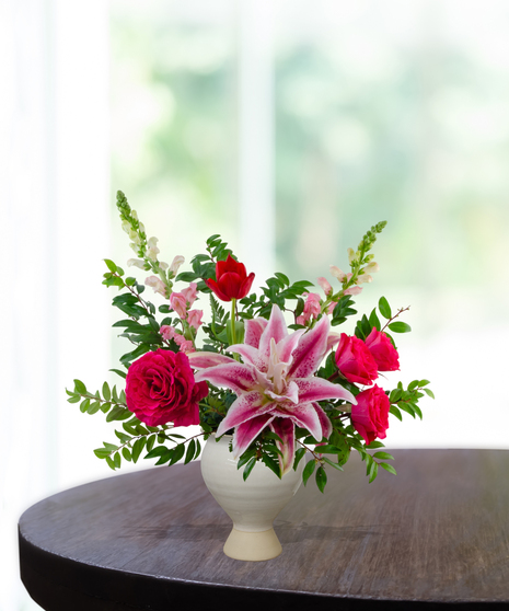 Legally Pink flower arrangement featuring hot pink roses, pink lilies, pink tulips, and white veronica in a matte cream ceramic pedestal vase.